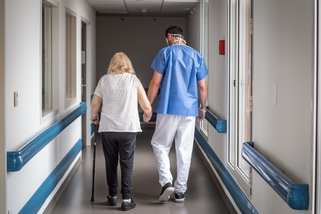 Photo représentant une patiente en train de marcher avec un professionnel de santé qui lui tient la main, dans les couleurs de la clinique des minimes